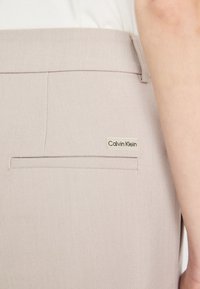 Beige op maat gemaakte broek van textuurstof met een achterzak; heeft een Calvin Klein logo-label nabij de zak.