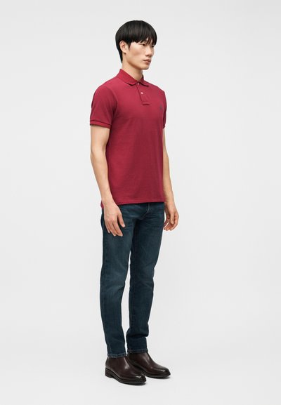 Νεαρός άνδρας όρθιος, φοράει κόκκινο πουκάμισο πόλο, σκούρο μπλε τζιν και σκού ρα καφέ μποτάκια μέχρι τον αστράγαλο, με φόντο ένα απλό ανοιχτό χρώμα.