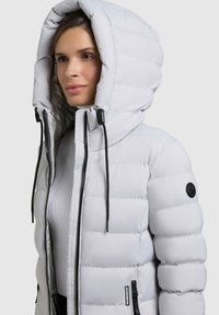 Hellgraue Steppjacke mit großer Kapuze, schwarzem Zubehör und horizontalem Steppmuster. Mit Frontreißverschluss und seitlichen Taschen.