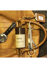 AHAVA Soothing After-Shave Moisturizer i en beige behållare, bredvid en silver rakkniv och en svart klocka på en brun läderväska.