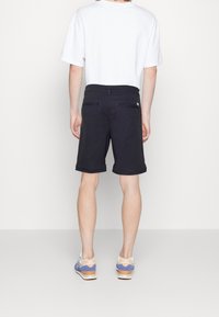 Person i svarta knälånga shorts, vit t-shirt och blå och vita New Balance-sneakers, som står med ryggen mot en enhetlig bakgrund.