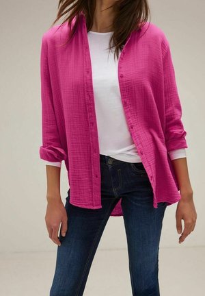 Frau, die ein hellpinkes Hemd über einem weißen Langarmshirt und dunkelblaue Jeans trägt, vor einem schlichten Hintergrund stehend.