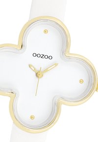 Montre avec bracelet en silicone blanc, dotée d'un boîtier en forme de trèfle avec des bords dorés et un cadran blanc. Logo "OOZOO" en noir et aiguilles dorées.