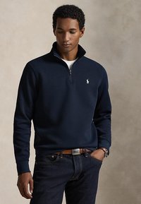 Muž nosící tmavě modrou mikinu na zip do půlky se světle modrým logem polo, tmavé džíny, hnědý pásek a náramkové hodinky, dívá se dolů na jednobarevném pozadí.