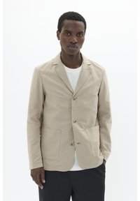 Beige cotton blazer with a lapel collar, three buttons, and front pockets, worn over a white shirt. Smooth texture, casual design.