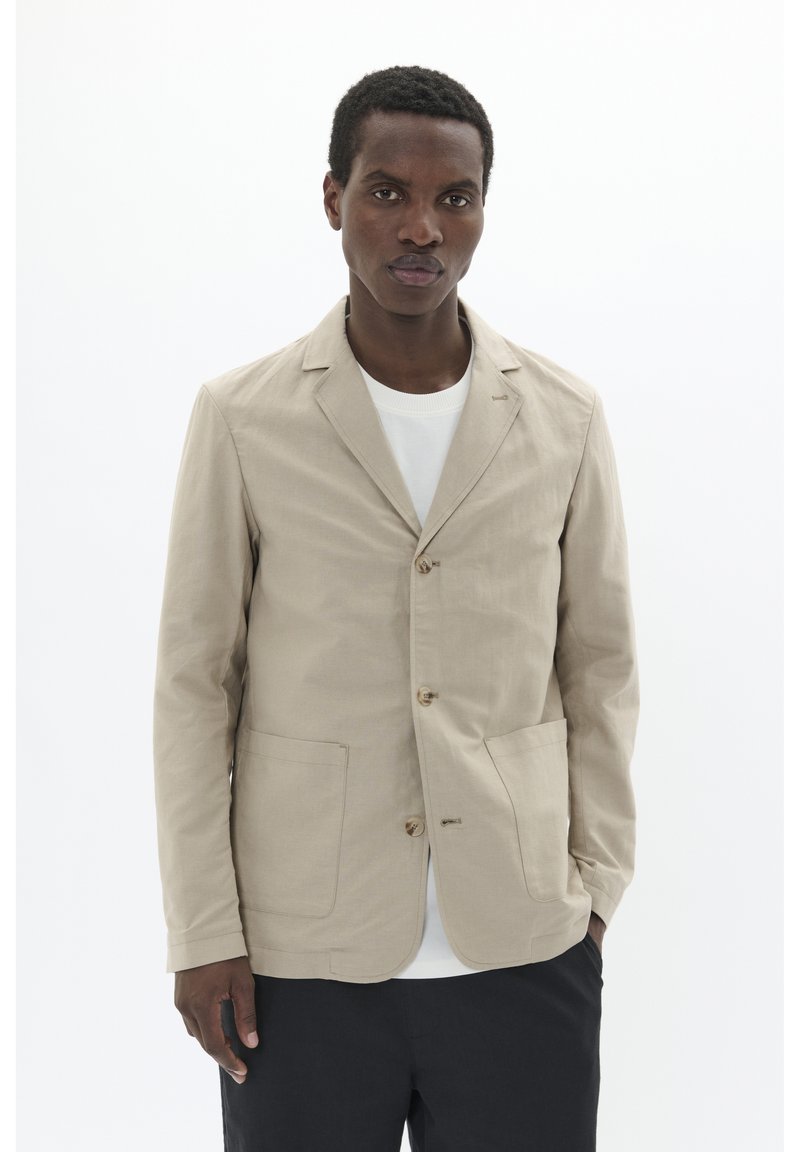 Beige cotton blazer with a lapel collar, three buttons, and front pockets, worn over a white shirt. Smooth texture, casual design.