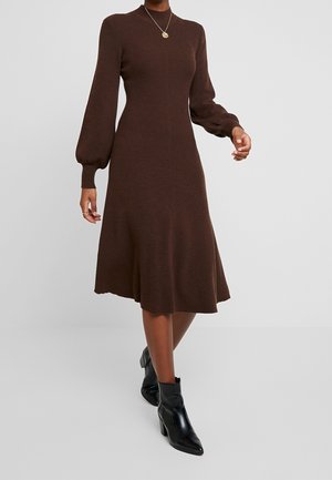 Vrouw draagt een bruine midi-jurk van gebreide stof met lange pofmouwen, zwarte enkellaarsjes en een gouden hangerketting tegen een effen achtergrond.