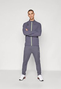 Siva športna jakna z zadrgo spredaj in neonsko zelenim poudarkom. Ujemajoče sive hlače in bele športne copate. Gladek tekstura tkanine.