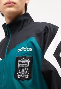 Person iført teal og sort Adidas-jakke med Liverpool Football Club-logo på brystet og hvide striber på ærmet.