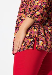 Blusa con stampa floreale in vivaci tonalità di rosa, arancione e nero, caratterizzata da una vestibilità ampia e fessure laterali, abbinata a pantaloni rossi elastici.