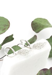 Silverhänge i form av ringar med intrikat bladformad filigrandesign, vilande på en vit stonyta omgiven av gröna blad.