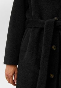 Zwarte wollen jas met een gestructureerde afwerking, voorzien van een ceintuur in de taille, grote knopen en zijzakken. Close-up weergave.