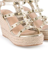 Sandalias de cuña doradas con tiras decoradas con tachuelas en forma de pirámide, suela de esparto trenzada y plantilla suave color beige.