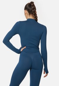 Top deportivo de manga larga en azul profundo, diseño ajustado, textura suave, con un cuello alto y agujeros para los pulgares en los puños. Combinado con leggings a juego.