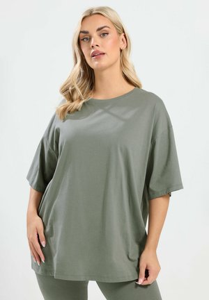 Mujer rubia vistiendo una camisa de manga corta y pantalones a juego de color verde oliva de corte holgado, de pie con las manos relajadas a los lados.