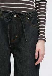 Melnas augsta vidukļa džinsa bikses ar redzamu jostu vidukļa joslā, kontrastējošu oranžu šuvumu un sānu kabatām. Izgatavotas no mīksta un teksturēta auduma.