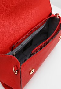 Borsa a tracolla in pelle rossa con una tasca con zip, hardware dorato e fodera interna grigia; presenta una etichetta con il logo all'interno.