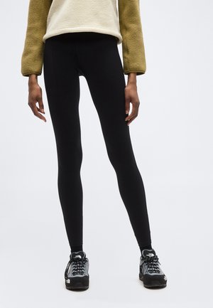 Osoba v čiernych legínach, béžovej flísovej bunde a čierno-sivých turistických topánkach stojaca na obyčajnom pozadí.