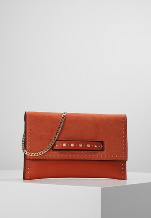 Pochette couleur rouille ornée de clous dorés, avec une découpe rectangulaire à l'avant et une bandoulière chaîne dorée, exposée sur un piédestal blanc.