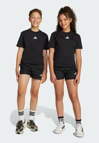 Čierne bavlnené tričká s bielym logom, skombinované s čiernymi šortkami. Jeden dieťa má zelené tenisky; druhé má biele tenisky s pruhmi.