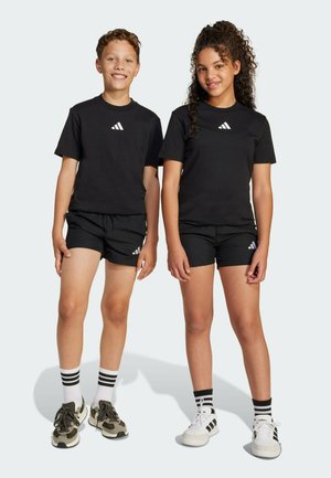 Camisetas de algodón negras con logo blanco, combinadas con pantalones cortos negros. Un niño lleva zapatillas verdes; el otro, zapatillas blancas con rayas.