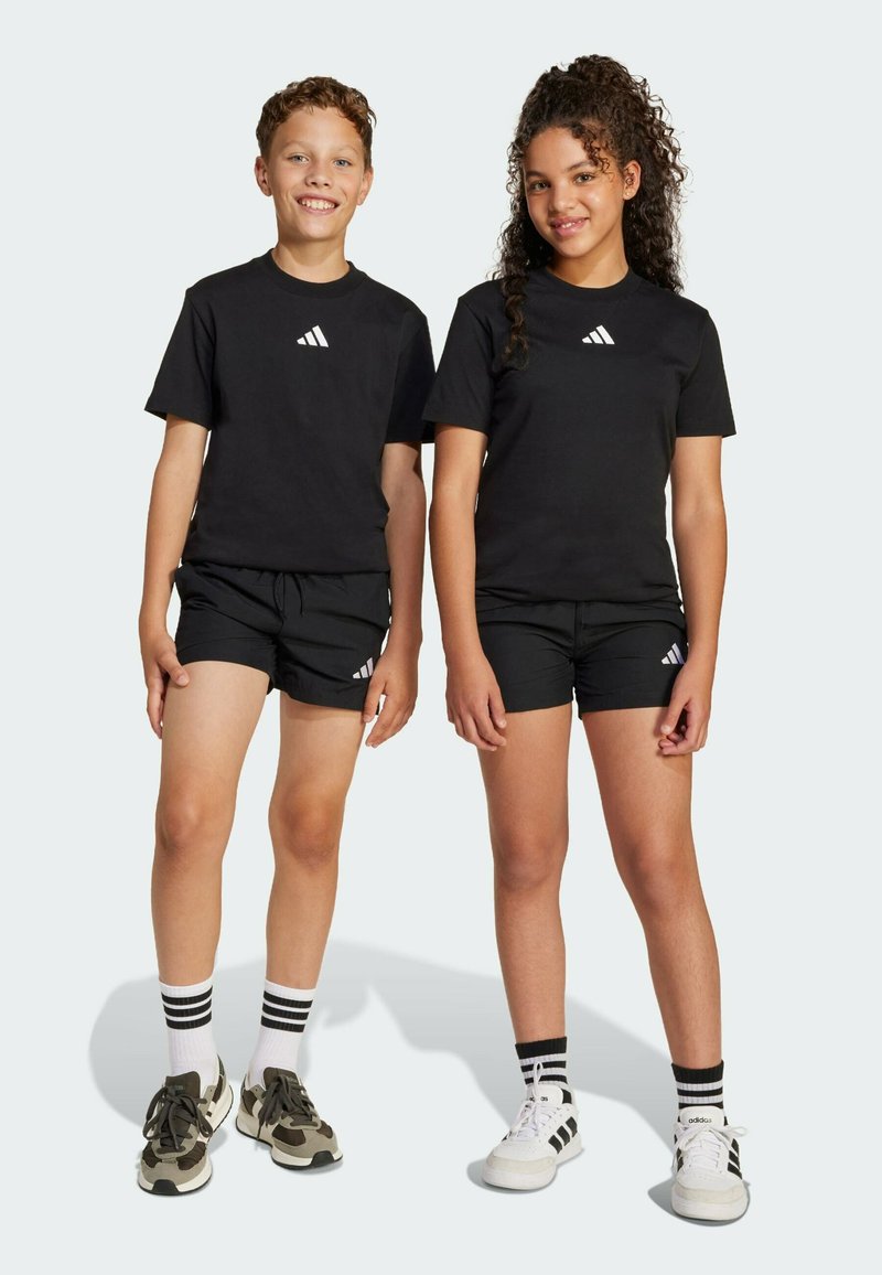 Čierne bavlnené tričká s bielym logom, skombinované s čiernymi šortkami. Jeden dieťa má zelené tenisky; druhé má biele tenisky s pruhmi.