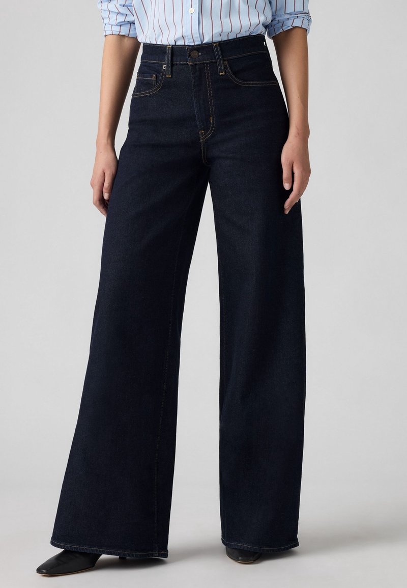Personne portant un jean taille haute, large et sombre, associé à des chaussures noires et une chemise bleu clair à rayures avec les manches retroussées.