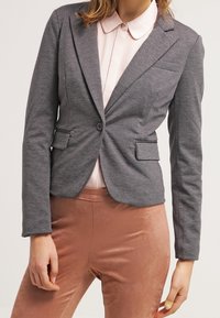 Blazer gris cintré avec deux poches avant et une fermeture à un bouton, porté sur une chemise à col rose clair et un pantalon couleur pêche.