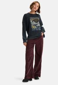 Grijs sweatshirt met lange mouwen en een grafisch ontwerp van bergen en bloemen. Gecombineerd met bordeauxkleurige corduroy broek met wijde pijpen en zwarte laarzen.