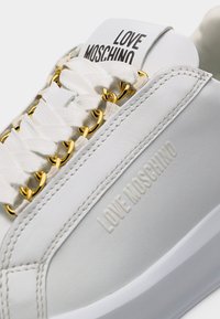 Gros plan sur une basket Love Moschino blanche avec des œillets en chaîne dorée et le nom de la marque embossé sur le côté.
