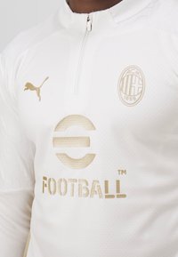 Weißes Fußballtrikot mit Halbreißverschluss-Design, goldenen Akzenten, Puma-Logo und gesticktem Teamlogo mit "Football" in fetter Schrift.