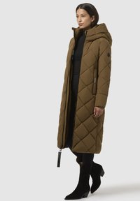 Lunga giacca impermeabile trapuntata verde oliva con cappuccio, dotata di zip frontale, tasche laterali e etichetta di stoffa. Indossata sopra abiti neri e stivali.