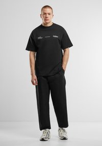 Tricou negru din bumbac, cu mâneci scurte, care prezintă un text imprimat alb. Combinat cu o pereche de pantaloni negri, largi și adidași sport multicolori.