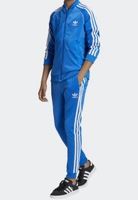 Modrý teplákový komplet z lesklého materiálu, se bílými pruhy na rukávech a kalhotách, doplněný černými teniskami.