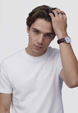 Mlad moški v beli majici se dotika las, na levi roki nosi uro z belim številčnico in temnim usnjenim trakom.