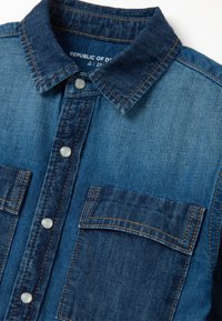 Camicia di denim con sfumatura di colore blu, caratterizzata da un colletto a punta, bottoni a scatto e due tasche sul petto con dettagli di cucitura.