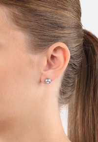 Silverfärgad metallstiftörhänge visat på ett öra. Rund form med en polerad, reflekterande yta som framhäver en slät textur.