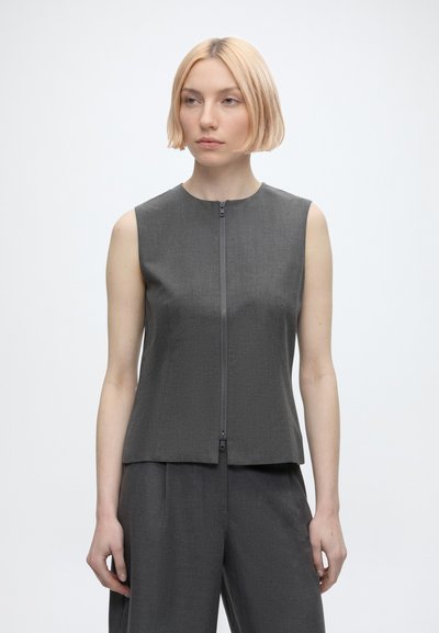 Femme aux cheveux blonds courts portant un top gris sans manches zippé et un pantalon large assorti sur un fond blanc uni.