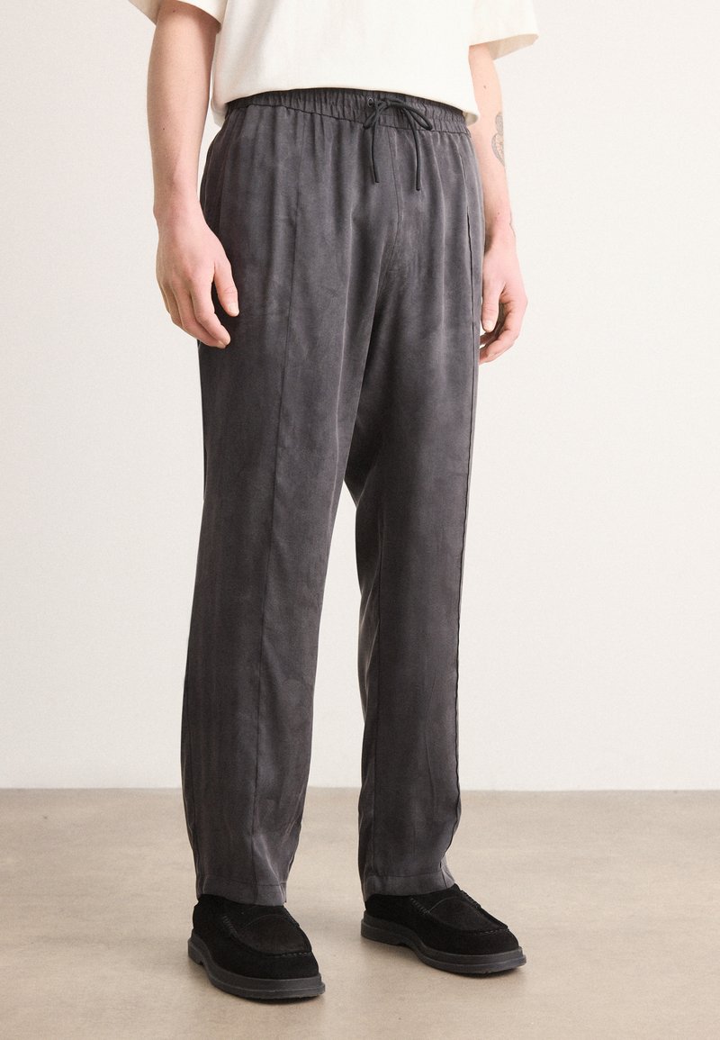 Pantalons gris, légers, à coupe décontractée avec taille élastique et cordon de serrage. Texture lisse, design à jambe droite, portés avec des chaussures noires.