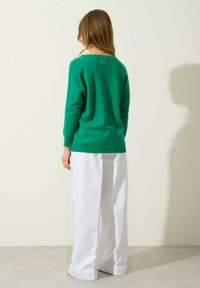 Grüner Strickpullover mit lockerem Schnitt und rundem Halsausschnitt, kombiniert mit weißen weiter geschnittenen Hosen, von hinten vor einem neutralen Hintergrund gezeigt.