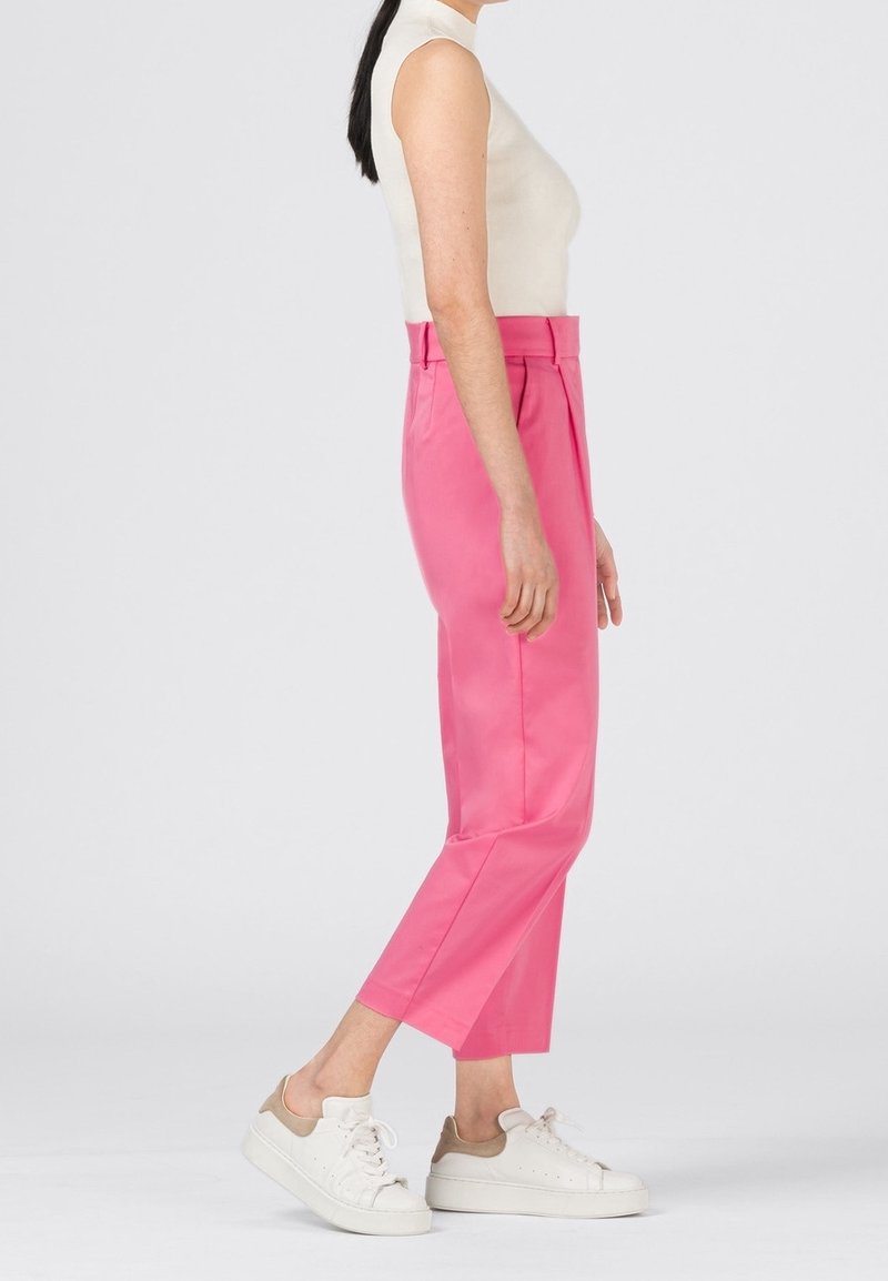 Femme aux cheveux noirs, debout de profil, portant un haut blanc sans manches, un pantalon rose taille haute et des baskets blanches sur un fond clair.