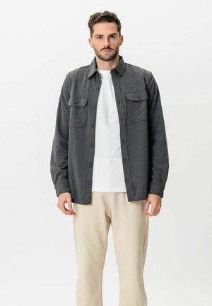 Muškarac odjeven u tamnosivu košulju-jaknu s gumbima preko bijele majice i bež hlača, stoji na jednostavnoj svijetloj pozadini.