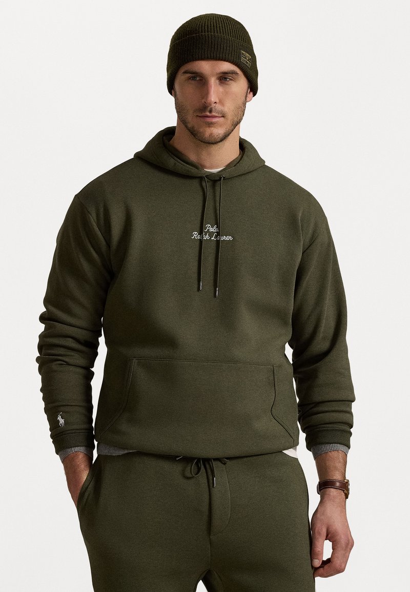 Ελιάς πράσινη hoodie από μαλακό ύφασμα με τσέπη καγκουρό, ρυθμιζόμενη κορδόνι και κεντητό λογότυπο; σε συνδυασμό με αντίστοιχες φόρμες και καφέ ρολόι χειρός.