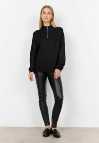 Zwarte geribde pullover met een hoge kraag en een halve rits aan de voorkant. Gecombineerd met strakke zwarte leggings en loafers. Gladde textuur.