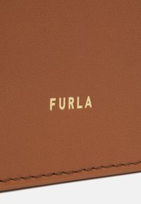 Superficie in cuoio tan con una finitura testurizzata, caratterizzata dal logo "FURLA" in rilievo dorato e cuciture visibili lungo i bordi.