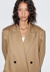 Tan double-breasted blazer with a textured fabric, notched lapels, and dark buttons, worn over a white top, against a plain background.