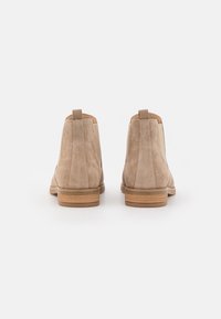 Beżowe zamszowe botki za kostkę o gładkim wykończeniu, minimalnych detalach szycia i z niskim drewnianym obcasem. Z tyłu pętle do łatwego zakładania.