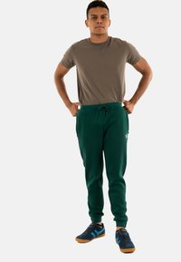 Pantalones jogger verde oscuro con bolsillos, combinados con una camiseta marrón claro y zapatillas negras con acentos azules y suelas de goma.