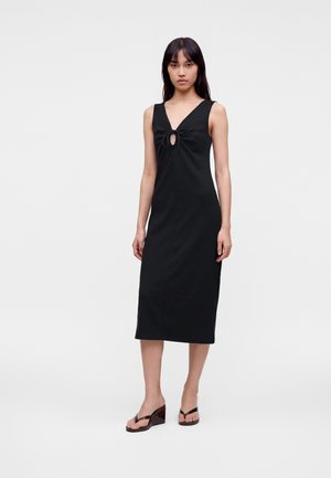 Jeune femme portant une robe midi noire sans manches avec un détail découpé à l'avant et des sandales compensées noires, debout devant un fond uni.