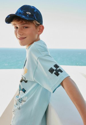 Hymyilevä poika, jolla on sininen, salaman kuvioitu lippalakki ja vaaleansininen graafinen t-paita, nojaa valkoiseen parvekkeeseen merimaiseman edessä.